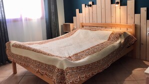 A bed with a patterned bedspread, a wooden headboard with built-in lighting, and a nightstand with a lamp. - Gîte de Leni (Torreilles)