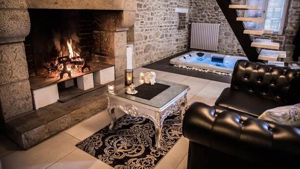 Une cheminée en pierre avec un feu de bois, un canapé en cuir noir, une table basse en verre et un mur en pierre.