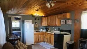 A compact kitchen with wooden cabinets, a refrigerator, a stove, and a microwave. There is a dining area with a chair and a table, and a small bookshelf with books. - Tiny House on the Farm: Watch Farm Animals, Near Buffalo River (Western Grove)