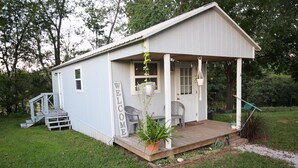 A small, single-story building with a porch, a welcome sign, and two chairs. - Tiny House on the Farm: Watch Farm Animals, Near Buffalo River (Western Grove)