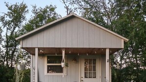 A small, single-story wooden structure with a porch, two chairs, a potted plant, and a hanging planter. - Tiny House on the Farm: Watch Farm Animals, Near Buffalo River (Western Grove)