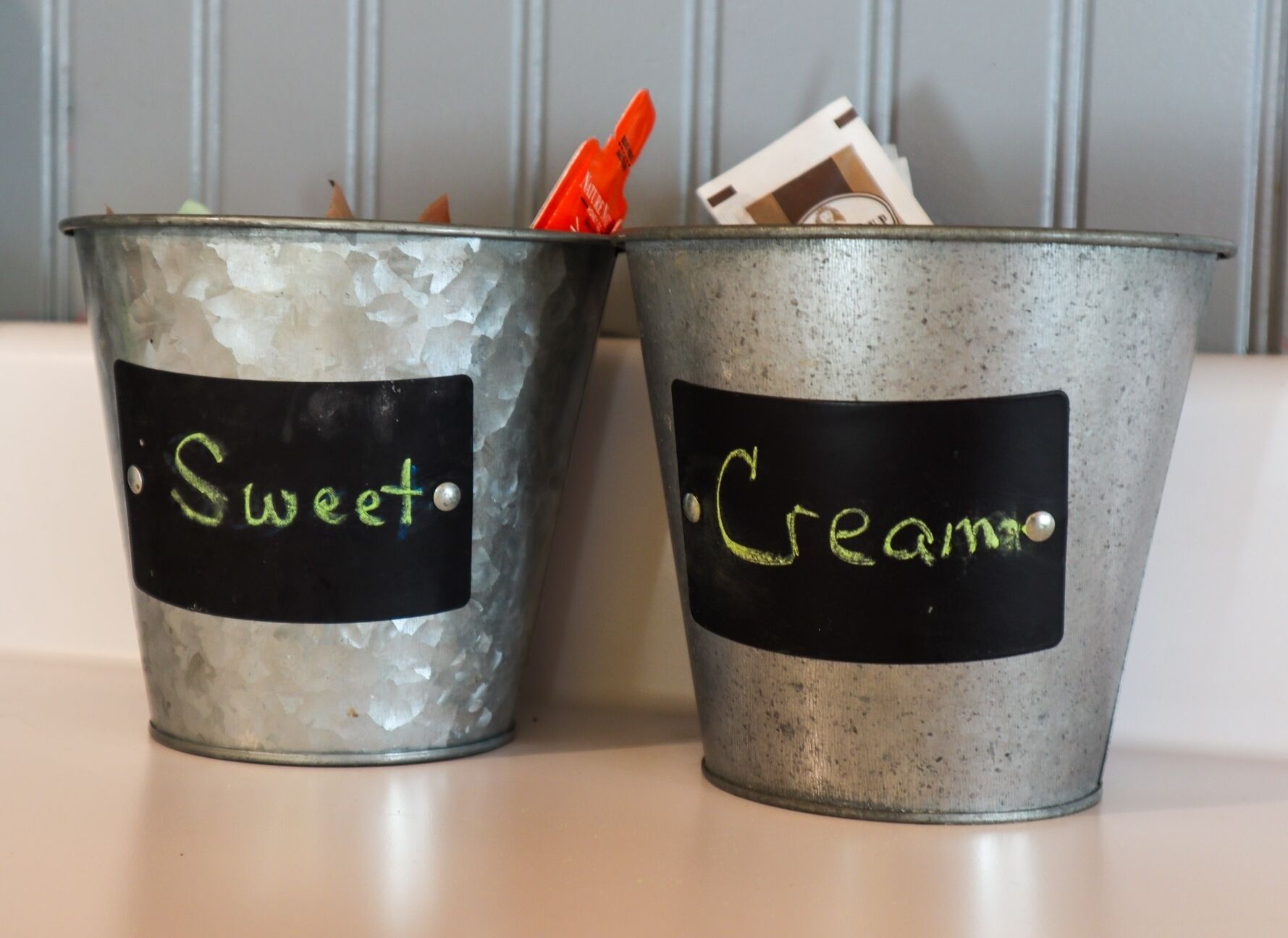 Due secchi metallici con le etichette 'Sweet' e 'Cream', e un'etichetta a lavagna, posati su una superficie.