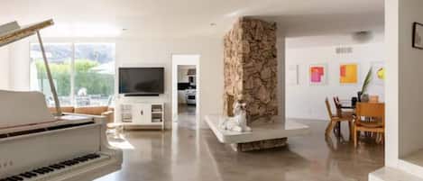 Amplia sala de estar con un piano, chimenea de piedra, televisor y comedor con sillas y mesa.