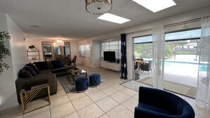 Amplia sala de estar con sofá, mesa de centro y otomanas. Se ve un televisor y un comedor.