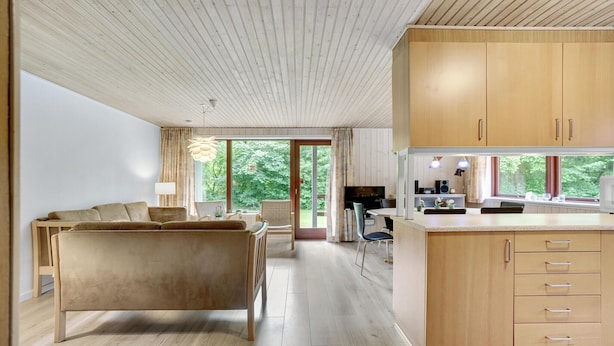 A modern living room with a sofa, a dining area with a table and chairs, a kitchen with wooden cabinets and a window, and a ceiling with wooden slats. - 4 bedroom beautiful home in Hemmet (Hemmet)