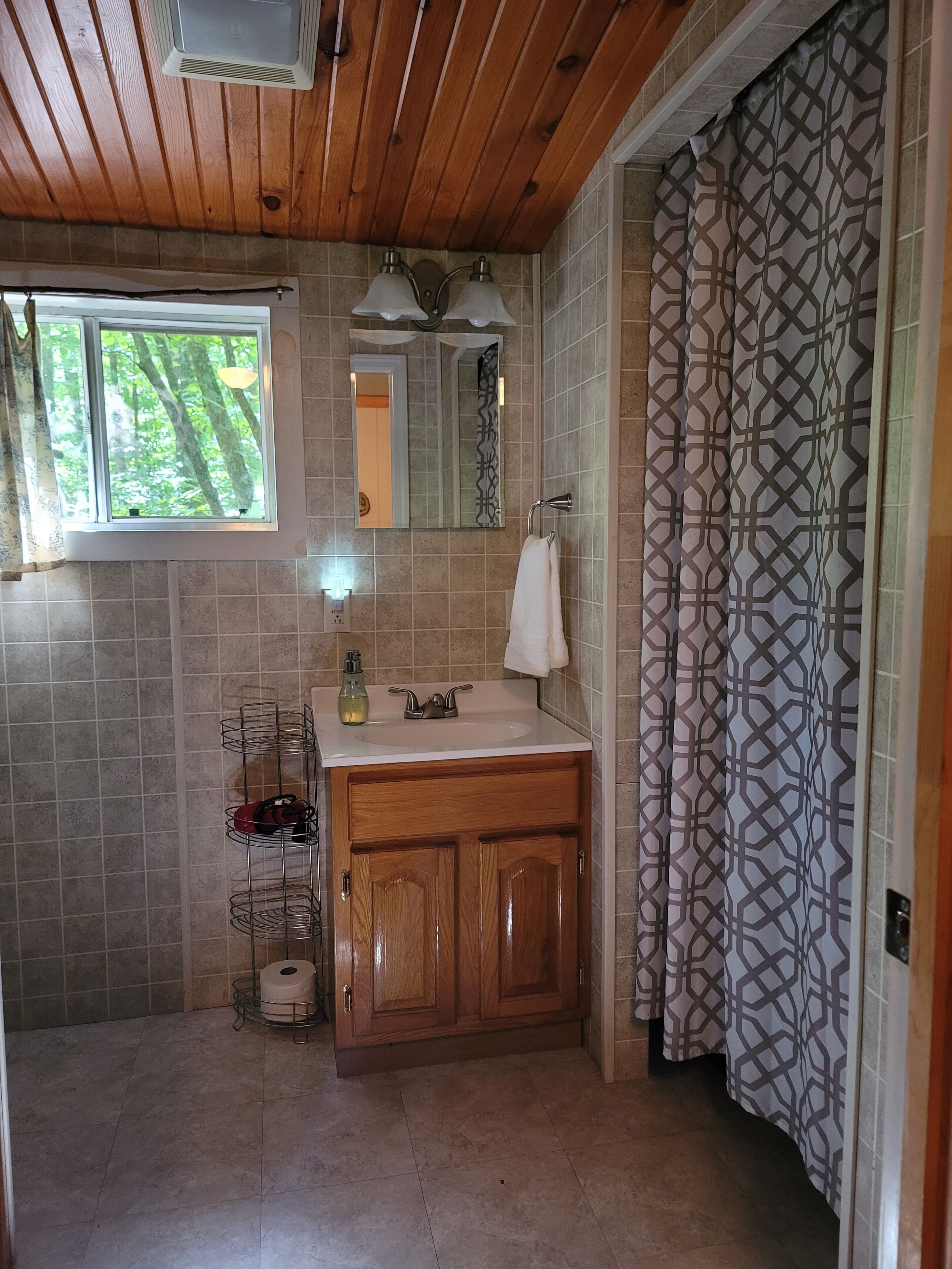 A bathroom with a wooden ceiling, a window with a view of trees, a sink with a towel, and a patterned curtain.
