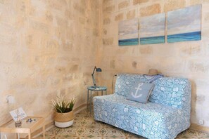 A room with a patterned sofa, a small side table, a lamp, a potted plant, and a wall with a painting.