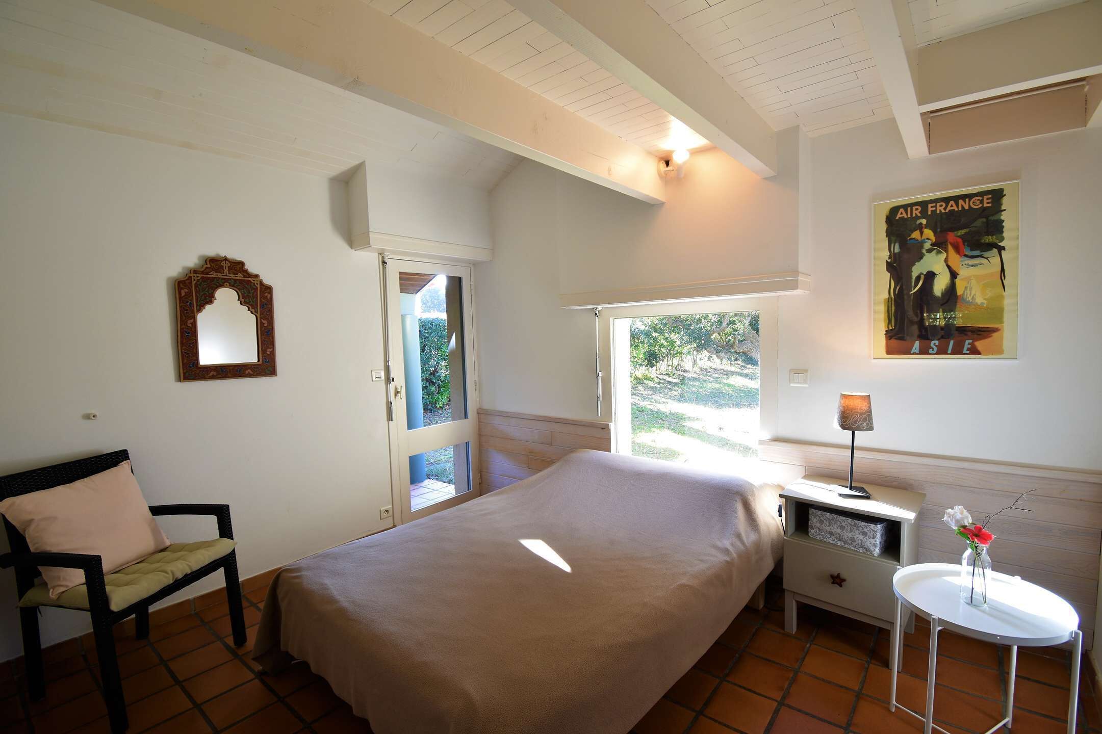 Une chambre à coucher avec un lit, une chaise, une table de chevet avec une lampe, une petite table avec des fleurs, une affiche au mur et une fenêtre donnant sur un paysage verdoyant.
