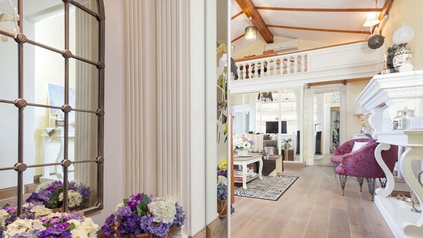 A spacious living area with a high ceiling, wooden beams, and a staircase leading to an upper level. There are two large windows with decorative iron grilles, and the room is furnished with a purple armchair, a white console table, and a mirror reflecting the room. - Winter Garden - NAVONA SQUARE (Roma)