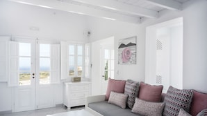 Een lichte, witte kamer met een grijze bank, een witte salontafel en uitzicht op de zee door grote ramen.