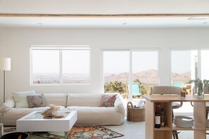 A spacious living room with a large sofa, a coffee table, and a dining area with a table and chairs. There are large windows offering a view of the mountains. - Minimalist desert retreat (Joshua Tree)