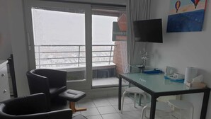 Een kamer met een glazen tafel, twee zwarte stoelen, een witte stoel, een televisie aan de muur en een schilderij van luchtballonnen.