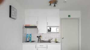 Een kleine kitchenette met witte kastjes, een magnetron, een spoelbak en een kookplaat. Er hangt een lijstje aan de muur en boven de magnetron is een klein raam.