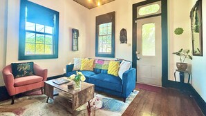 Un salon avec un canapé bleu, un fauteuil rose et une table basse en bois. Il y a de grandes fenêtres, une porte avec un panneau de verre et une plante suspendue.