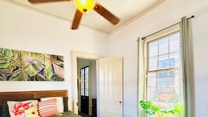 Une chambre à coucher avec un lit, un ventilateur de plafond, une fenêtre avec des rideaux et une plante sur une table de chevet.