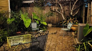 Une terrasse extérieure avec un sol en briques, des chaises en métal, une table en verre et des guirlandes lumineuses.