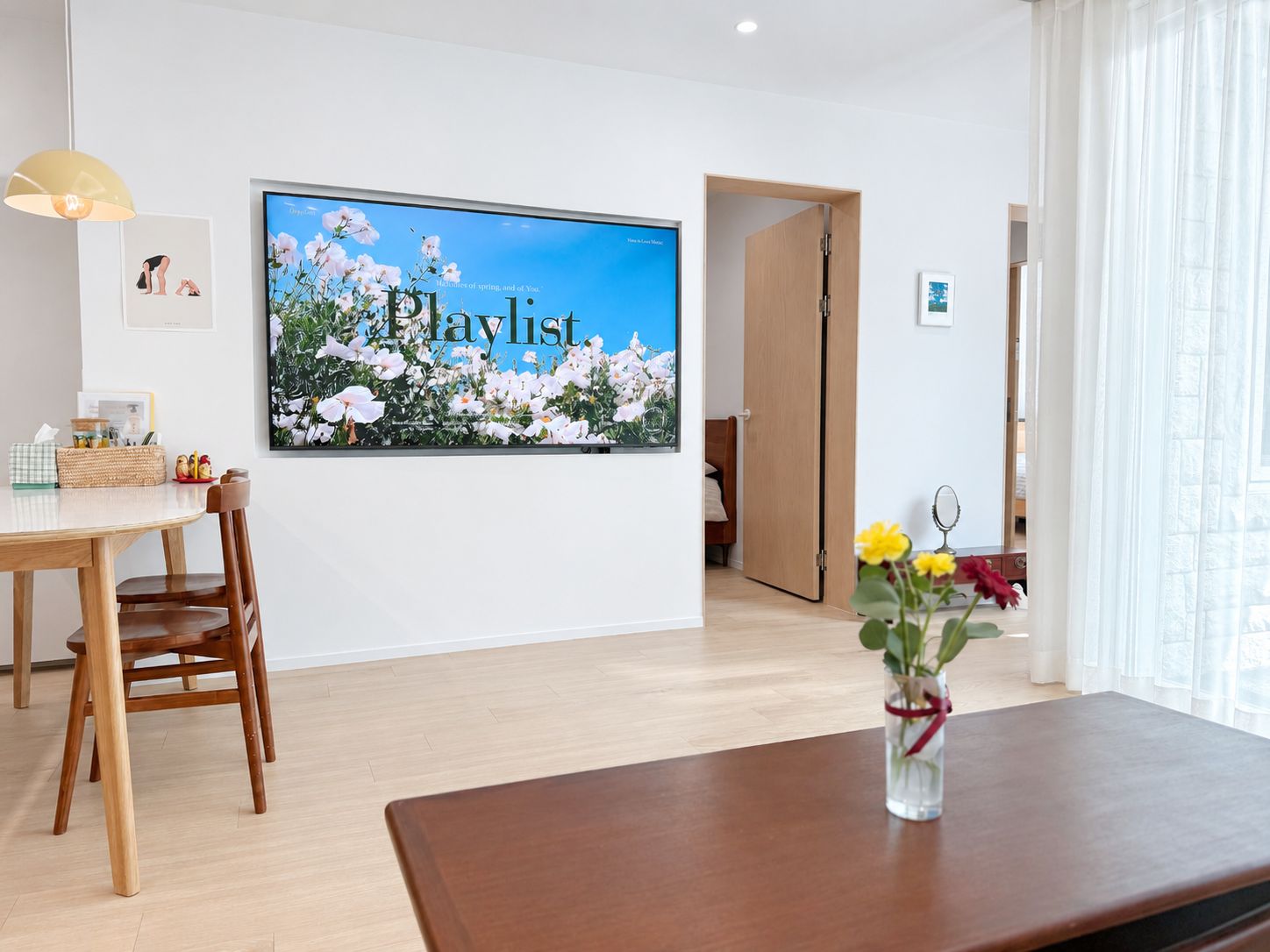A modern room with a large flat-screen TV displaying a floral image, a wooden dining table with chairs, and a vase with flowers on a wooden table.