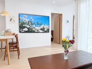 A modern room with a large flat-screen TV displaying a floral image, a wooden dining table with chairs, and a vase with flowers on a wooden table. - 6Min Hongdae | 83㎡ Family House | Private Yard (서울)