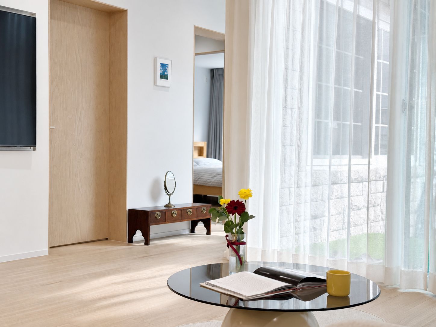 A modern hotel room with a round glass table, a wooden dresser, a bed, and a large window with sheer curtains.