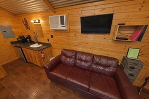 Interior de una cabaña de madera con kitchenette, un sofá de cuero, televisor y una pequeña estantería.