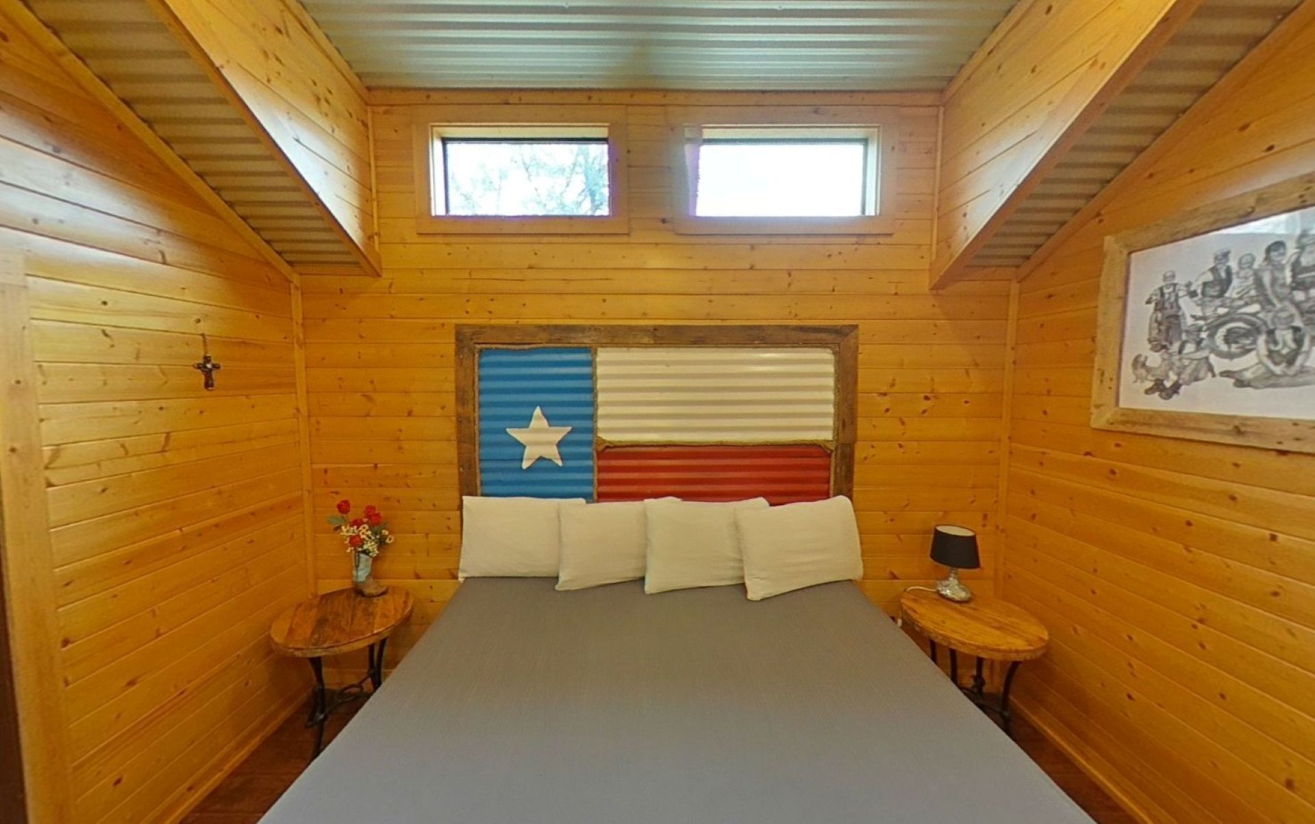 A room with wooden walls, a bed with white pillows, a small table with a vase of flowers, and a framed picture on the wall.
