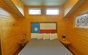 Una habitación con paredes de madera, una cama con almohadas blancas, una mesita con un jarrón de flores y un cuadro en la pared.