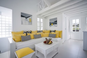 A bright, white interior with a sofa set adorned with yellow and patterned cushions, a coffee table with fruit, and artwork on the walls.