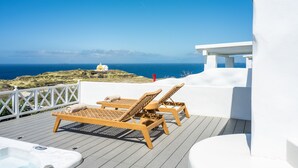 Una terrazza con due poltrone in legno e una vasca idromassaggio, con vista su un paesaggio costiero e un cielo sereno.
