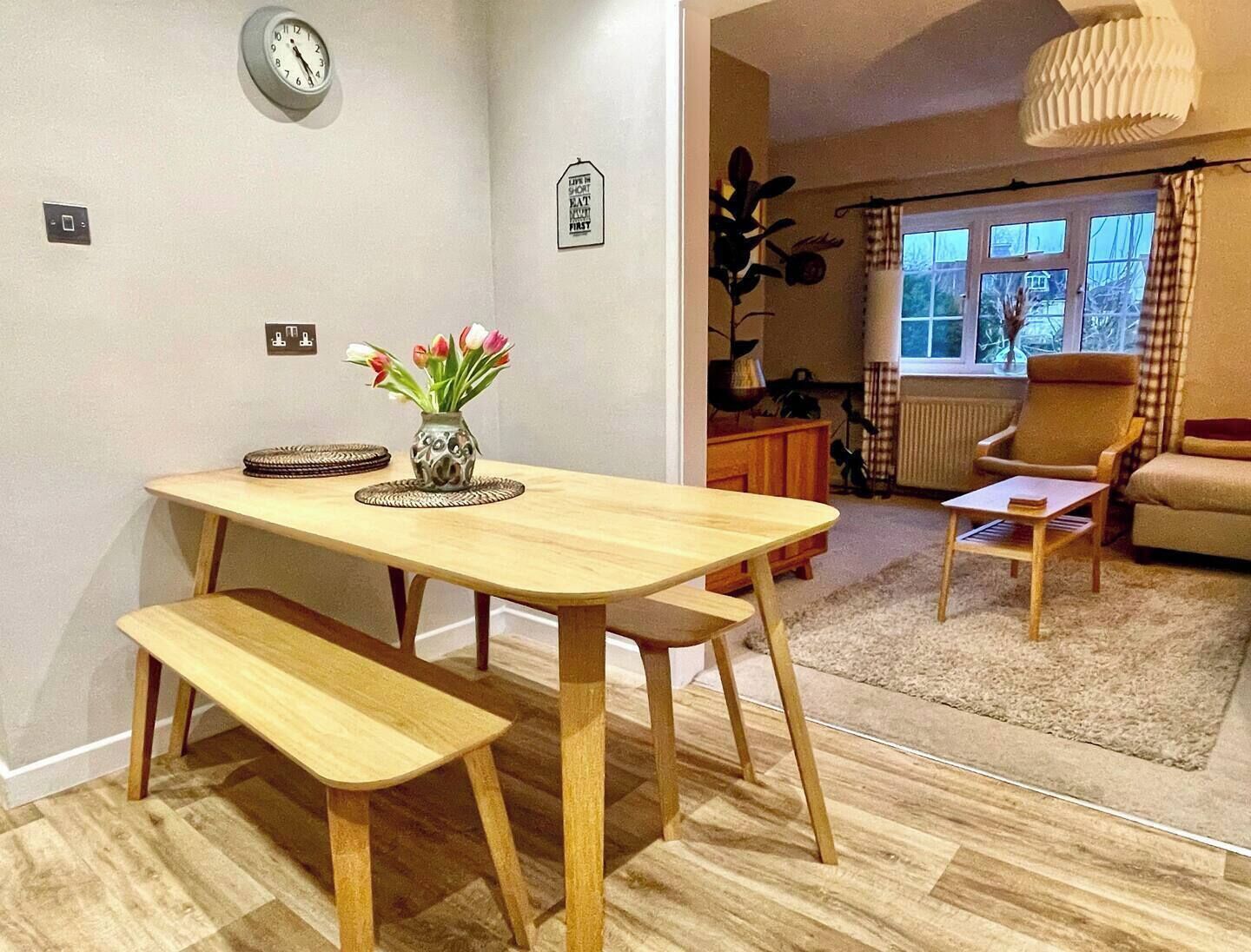 Ein Essbereich mit einem Holztisch und einer Bank, einer Vase mit Blumen sowie einer Uhr an der Wand.