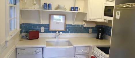 Cocina compacta con armarios blancos, lavavajillas, estufa y microondas. Se observa una ventana con persianas, un estante con platos y tazas, y un rollo de toallas de papel.