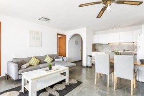 Uma sala de estar moderna com um sofá cinzento, uma mesa de centro branca e uma área de jantar com mesa e cadeiras de madeira.
