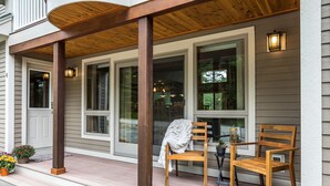 Une véranda en bois avec deux chaises et une table, un espace couvert doté d’un plafond en bois, et une porte avec une fenêtre.