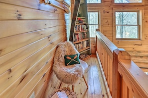 Een gezellig blokhutinterieur met houten wanden, een comfortabele bonten fauteuil, een boekenplank en een raam met uitzicht op bomen.