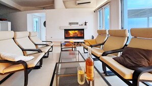 A modern living room with leather sofas, a glass coffee table, a fireplace, and a bottle of whisky. - Luxury Private Villa with Infinity Pool & Sunset Sea Views – Paros (Parikia-Paros)
