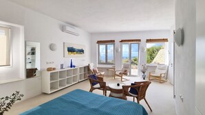A spacious room with a large bed, a dining area with wicker chairs, a white console table, and a view of the sea through large windows. - Luxury Private Villa with Infinity Pool & Sunset Sea Views – Paros (Parikia-Paros)