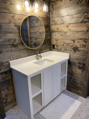 A bathroom with a white vanity, a round mirror with a gold frame, and three wall-mounted lights. - *Brand New* Sweet Pines Lodge: Modern Luxury nestled in on a private cul-de-sac  (Broken Bow)