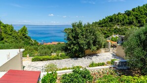 A coastal view with a stone courtyard, lush greenery, and a clear blue sea. - Vacation House Marija -Two Bedroom Holiday Home with Terrace and Sea View (Korcula)