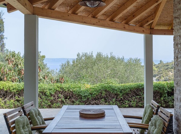 A covered outdoor seating area with a table and chairs, overlooking a body of water and greenery.