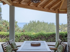 A covered outdoor seating area with a table and chairs, overlooking a body of water and greenery.