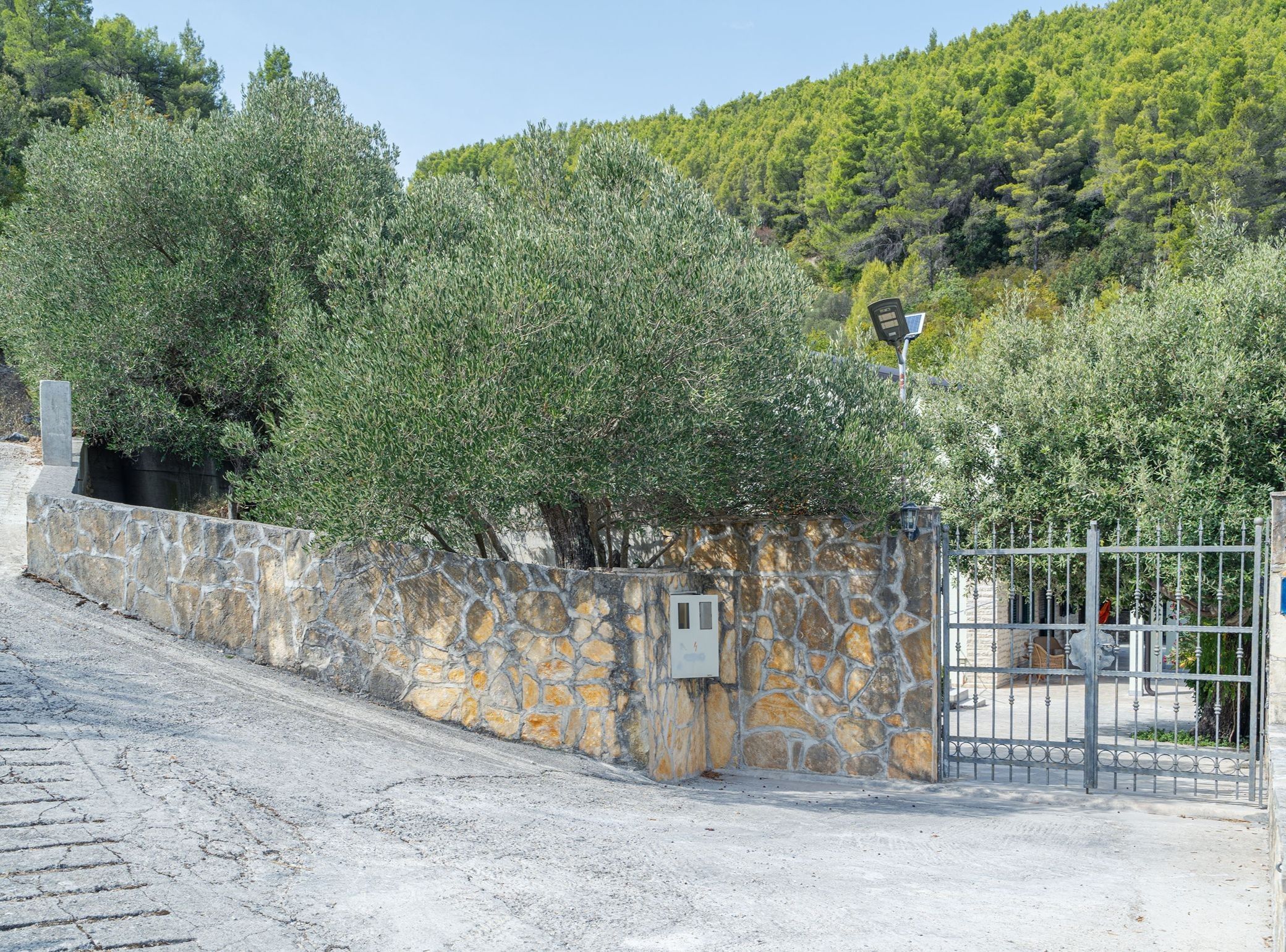 A stone wall with a metal gate, a paved path, and a hillside with trees.