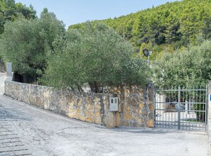 A stone wall with a metal gate, a paved path, and a hillside with trees.