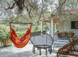 Un hamac et une chaise en osier sous un arbre, avec du mobilier d’extérieur et un bâtiment en arrière-plan.