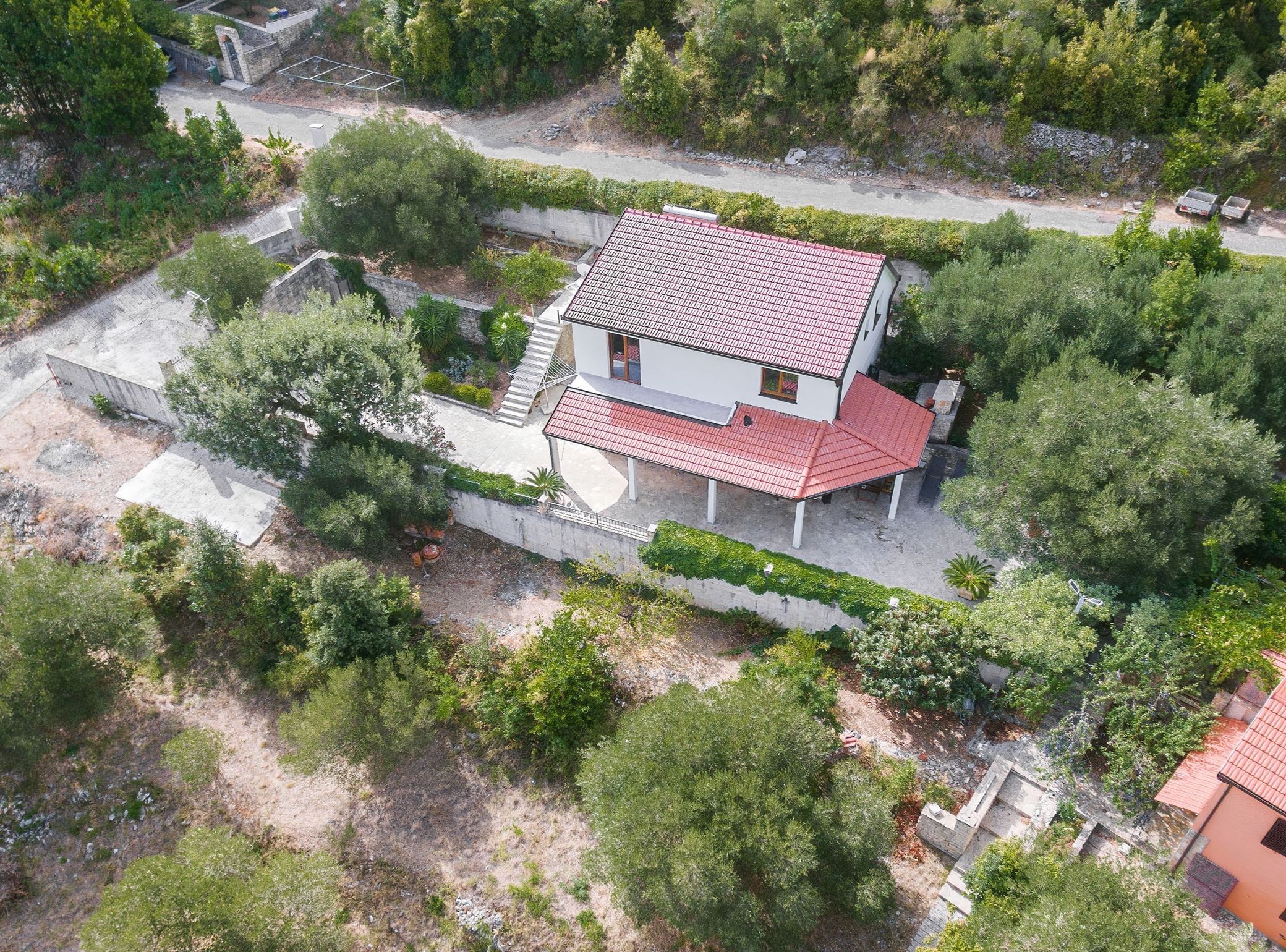 A two-story house with a red-tiled roof, surrounded by greenery and a paved driveway.