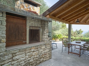 A stone building with a wooden door and window, outdoor seating area with tables and chairs, and a pergola with a hanging light fixture.