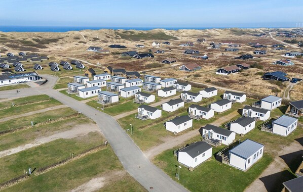 An aerial view of a caravan park with compact, white mobile homes arranged in rows, surrounded by grassy fields and dunes, with a clear view of the sea in the background.