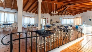 A spacious dining area with multiple tables and chairs, a kitchen area with a refrigerator and sink, and a wooden ceiling with exposed beams. - Vignola Baia dei Mirti B4 (Vignola Mare -SS-)