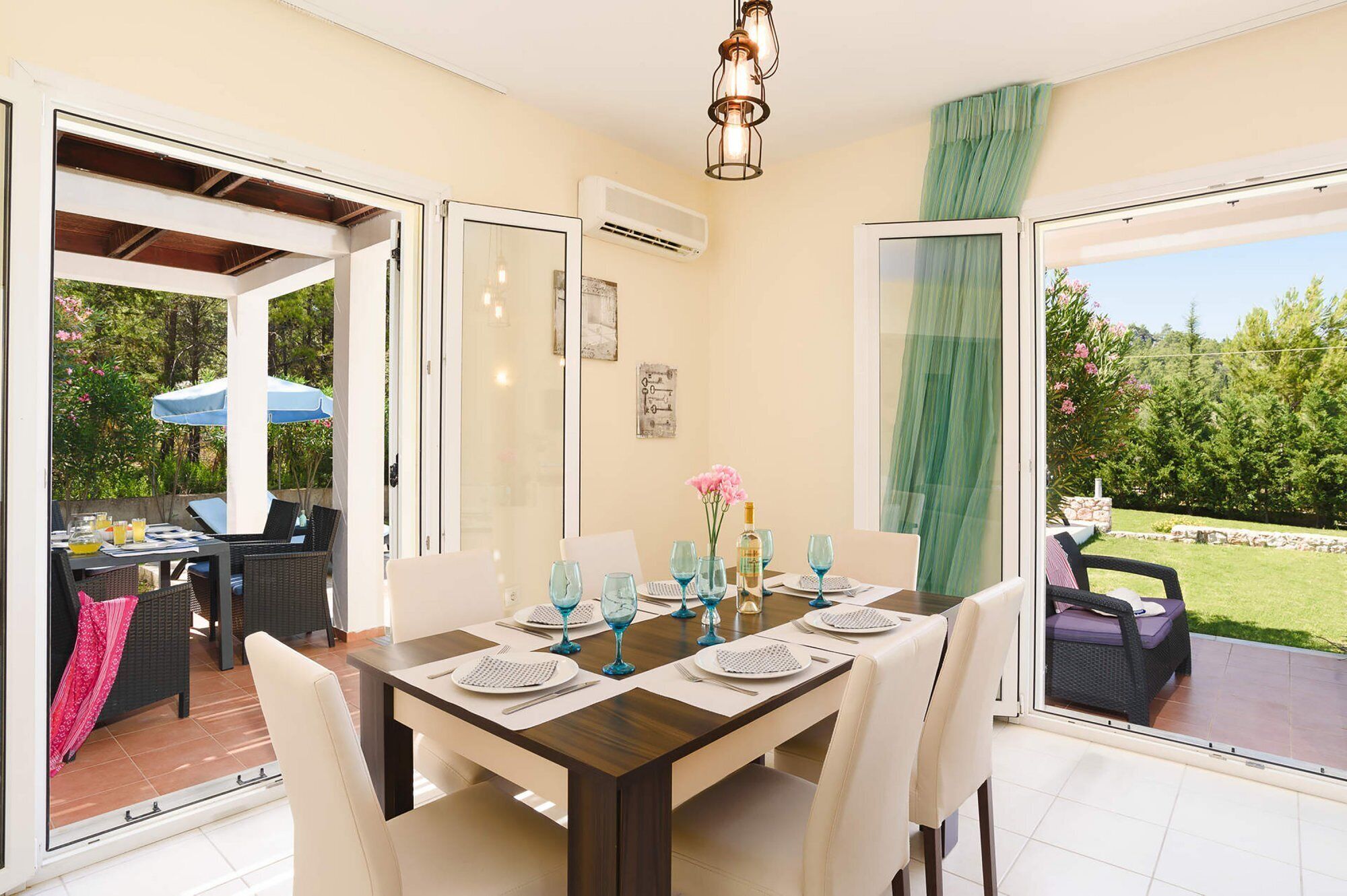 Un coin repas avec une table dressée pour quatre personnes, une vue sur le jardin à travers des portes vitrées et un luminaire au plafond.