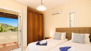 Ein Hotelzimmer mit zwei Betten, einem Holzschrank, einer Klimaanlage und einem Balkon mit Blick auf Grünflächen.
