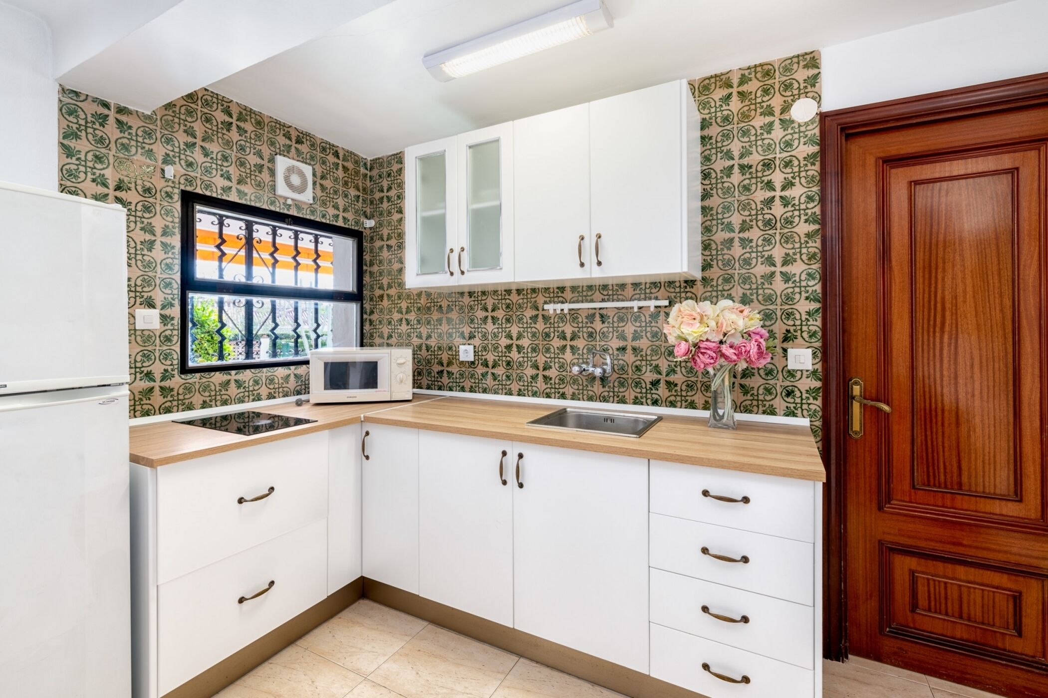 Una cocina con armarios blancos, un microondas y un lavamanos. Se observa una puerta de madera y una ventana con un marco decorativo.