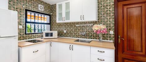 Una cocina con armarios blancos, un microondas y un lavamanos. Se observa una puerta de madera y una ventana con un marco decorativo.